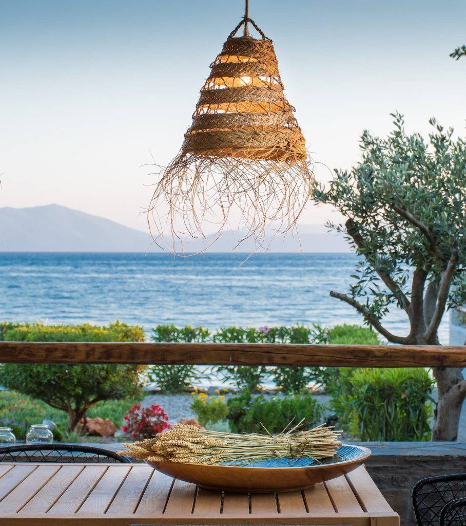 Seaside dining with a woven lamp, wooden table, and an olive tree
